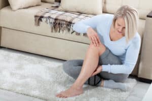 Middle-aged woman sitting on floor examining her lower leg for pain or skin changes related to vein issues